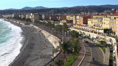 Turquoise waves crashing along the pebble beach of the Promenade des Anglais, with colorful buildings and palm trees lining the seafront - Starpik Stock