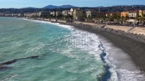 Turquoise waves crashing along the pebble beach of the Promenade des Anglais, with colorful buildings and palm trees lining the seafront - Starpik Stock