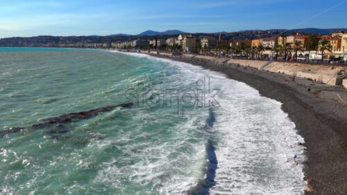 Turquoise waves crashing along the pebble beach of the Promenade des Anglais, with colorful buildings and palm trees lining the seafront - Starpik Stock