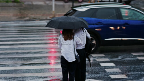 Two girls holding an umbrella waiting at a pedestrian crossing on a rainy day in Chisinau, Moldova - Starpik Stock