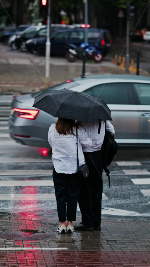 Two girls holding an umbrella waiting at a pedestrian crossing on a rainy day in Chisinau, Moldova. Vertical - Starpik Stock