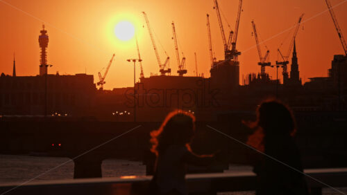 Two women standing by the railing watching a vibrant sunset over cranes and the London skyline, London, England - Starpik Stock