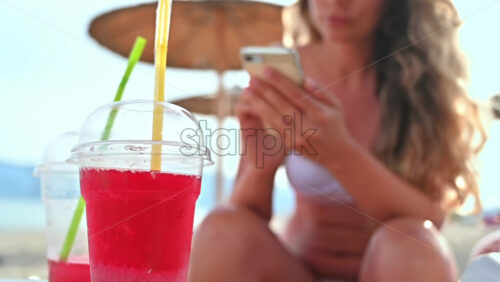 Video – A red cocktail on the beach. Caucasian woman with smartphone sitting on the sunbed on the background. Sea, slow motion - Starpik Stock