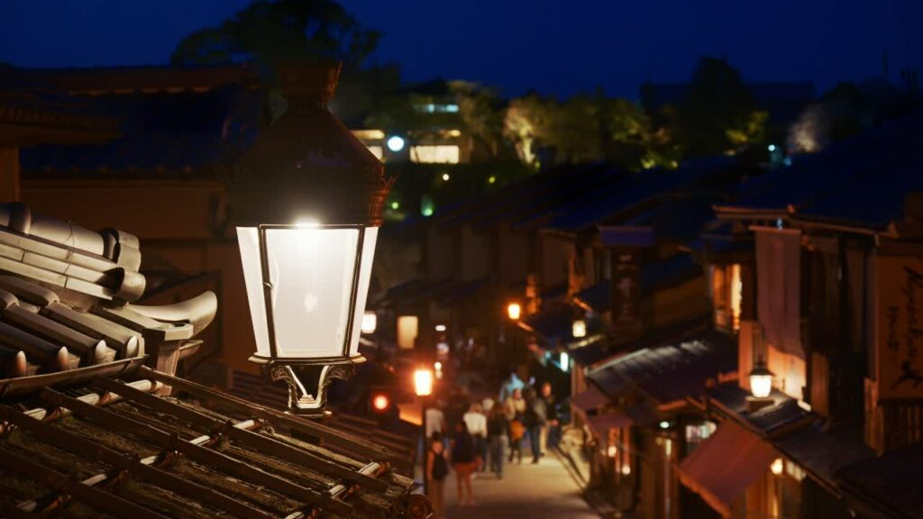 Video – Kyoto, Japan – April 11, 2025: Close up of a street lamp with a blurred view of people walking on the Ninenzaka stone-paved pedestrian road, lined with traditional buildings and shops in the evening - Starpik Stock