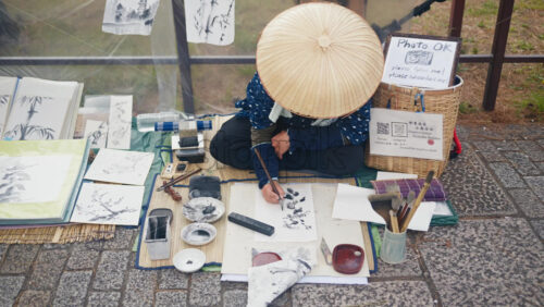 Video – Kyoto, Japan – April 11, 2025: Person wearing a traditional Japanese hat painting Sumi-e traditional style in black and white on the street of Gion district - Starpik Stock