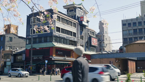 Video - Kyoto, Japan - April 12, 2025: People and cars moving on the streets of the Gion area in daylight. Translation: "Izutsu Yatsuhashi Honpo - Kyoto Gion Main Store"