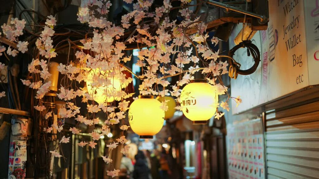 Video – Shinjuku, Japan – April 12, 2025: Lighted paper lanterns hanging with cherry blossom flower decorations on the on the Omoide Yokocho Memory Lane in the evening. Translation: “Restaurant name” - Starpik Stock