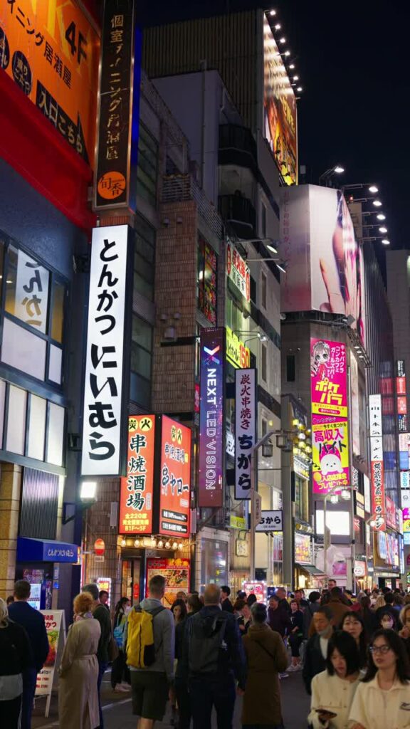 Video – Tokyo, Japan – April 12, 2025: View of the vibrant life of Shinjuku City with people moving on the streets with lighted buildings in the evening. Translation: “Company names”. Vertical - Starpik Stock