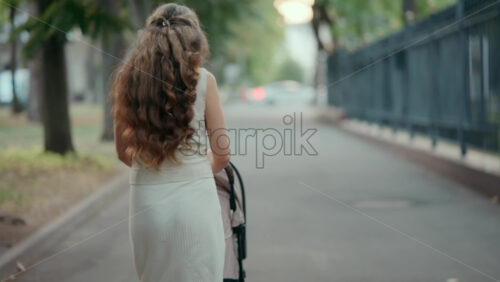 View from the back of a woman with long wavy hair pushing a stroller along a tree-lined park pathway - Starpik Stock