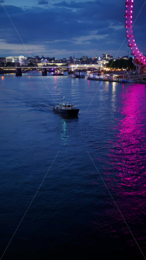 View of a boat moving on the river with the London Eye glowing pink and County Hall lit in green in the evening. Vertical - Starpik Stock