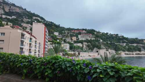 Villefranche-sur-Mer, France – July 18, 2025: People climbing a coastal pathway lined with rocky cliffs and lush flowering plants, leading towards pastel coloured apartment buildings - Starpik Stock