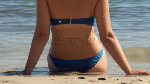 Woman in a blue bikini sitting at the waterline, arms braced behind her as small waves lap the beach - Starpik Stock