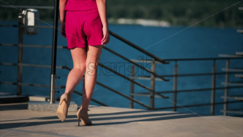 Woman in a hot pink, shorts suit with tan high heels walking on a scenic terrace with a view of a calm body of water - Starpik Stock