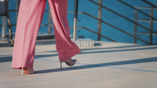 Woman in a pink suit with tan high heels walking on a scenic terrace with a view of a calm body of water - Starpik Stock