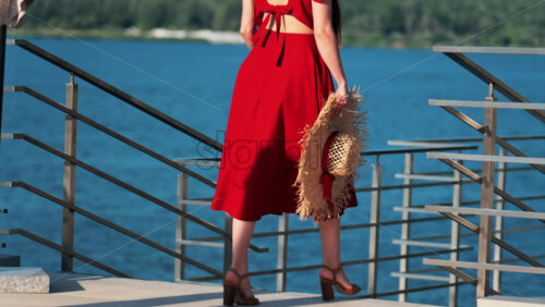 Woman in a red dress holding wide hat walking on a scenic terrace with a view of a calm body of water - Starpik Stock