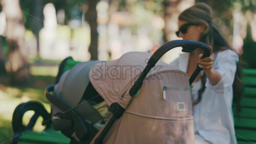 Young mother sitting on a park bench next to a stroller, enjoying a peaceful outdoor moment with her baby - Starpik Stock