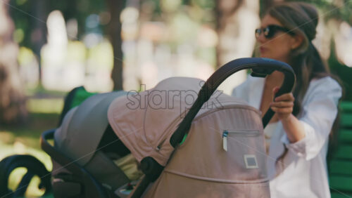 Young mother sitting on a park bench next to a stroller, enjoying a peaceful outdoor moment with her baby - Starpik Stock