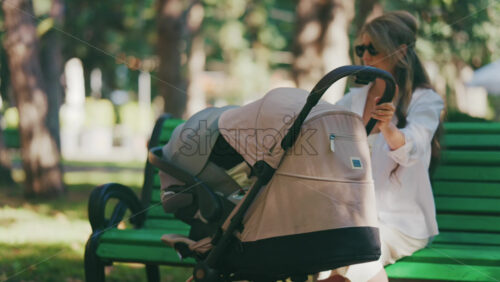 Young mother sitting on a park bench next to a stroller, enjoying a peaceful outdoor moment with her baby - Starpik Stock