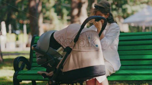 Young mother sitting on a park bench next to a stroller, enjoying a peaceful outdoor moment with her baby - Starpik Stock