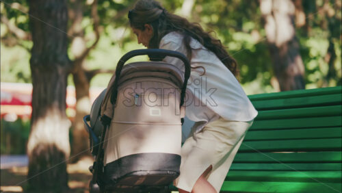Young mother sitting on a park bench next to a stroller, enjoying a peaceful outdoor moment with her baby - Starpik Stock