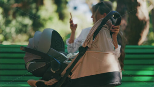Young mother sitting on a park bench next to a stroller, enjoying a peaceful outdoor moment with her baby - Starpik Stock