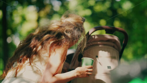 Young mother with long curly hair holding a coffee cup while leaning over a baby stroller in the park - Starpik Stock