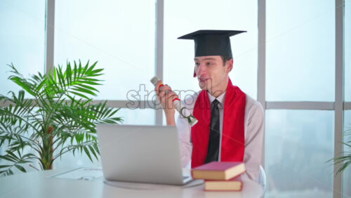 Video - Young man with graduation diploma talking on the laptop at home. Graduating online in virtual ceremony