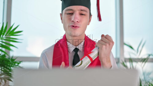 Video - Young man with graduation diploma talking on the laptop at home. Graduating online in virtual ceremony