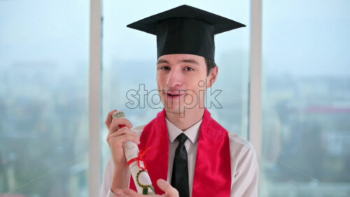 Video - Young man with graduation diploma talking towards the camera. Graduating online in virtual ceremony