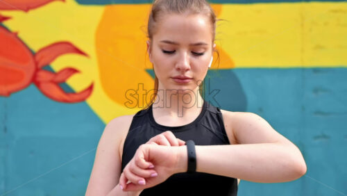 Video - Young blonde woman at outdoors training in sportswear is on her fitness bracelet, earphones, multicolored background. Slow motion