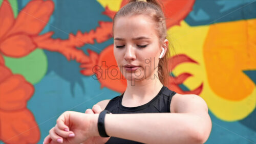 Video - Young blonde woman at outdoors training in sportswear is on her fitness bracelet, earphones, multicolored background. Slow motion