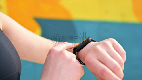 Video - Young woman at outdoors training in sportswear is on her fitness bracelet, earphones, multicolored background. Slow motion
