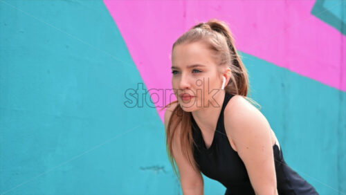 Video - Young focused blonde woman at outdoors training stopping to run, earphones, multicolored background. Slow motion