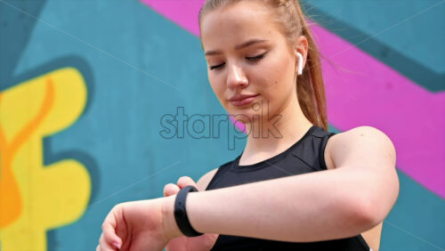 Video - Young blonde woman at outdoors training in sportswear is on her fitness bracelet, earphones, multicolored background. Slow motion