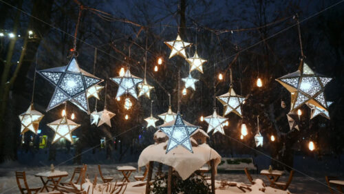Video - Wide shot of the star-shaped lanterns and coffee tables in the park during snowfall. Christmas decorations. Winter evening
