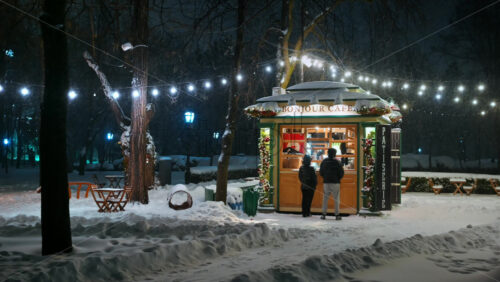 Video - Chisinau, Moldova - January 09, 2024: People buying coffee at the illuminated Bonjour coffee shop with Christmas decorations in the central park during snowfall. Winter evening