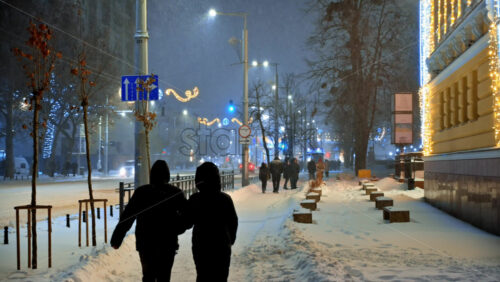 Video - Chisinau, Moldova - January 09, 2024: People walking on an illuminated street in the city center during snowfall in the evening. Christmas decorations. Blue hour