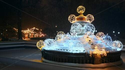Video - Chisinau, Moldova - January 09, 2024: Christmas decorations on the fountain in the central park during snowfall. Illuminated Bonjour coffee shop in background. Winter evening