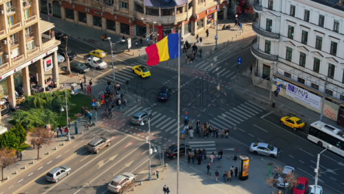 Video - Bucharest, Romania - March 03, 2024: Aerial drone view of a downtown intersection with moving traffic and walking people in sunlight. Waving national flag