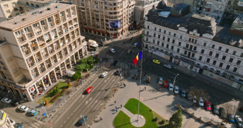Video - Bucharest, Romania - March 03, 2024: Aerial drone view of a downtown intersection with moving traffic and walking people in sunlight. Waving national flag