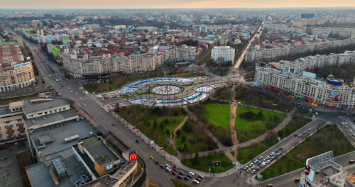Video - Bucharest, Romania - March 03, 2024: Aerial drone view of the city downtown at sunset. Roundabout intersection with moving cars, fountains and greenery. Palace of the Parliament in the distance