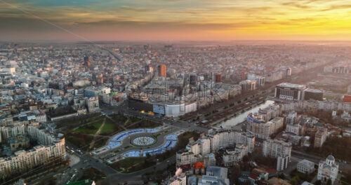 Video - Bucharest, Romania - March 03, 2024: Aerial drone view of the city downtown at sunset. Roundabout intersection with moving cars, fountains and greenery. Palace of the Parliament in the distance