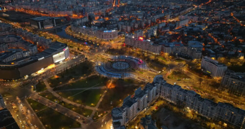 Video - Bucharest, Romania - March 03, 2024: Aerial drone view of the illuminated city downtown in the evening. Roundabout intersection with moving cars and fountains. Blue hour