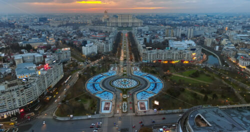 Video - Bucharest, Romania - March 03, 2024: Aerial drone view of the city downtown at sunset. Roundabout intersection with moving cars and fountains. Palace of the Parliament in the distance