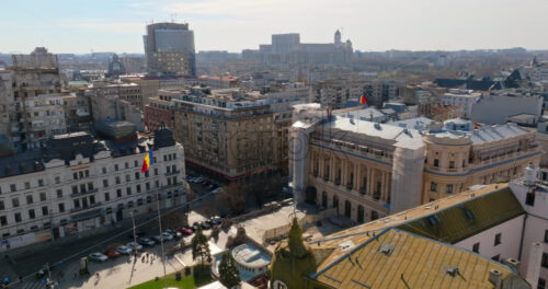 Video - Bucharest, Romania - March 03, 2024: Aerial drone view of a downtown road with moving traffic and walking people. Palace of the Parliament in background. Sunny day