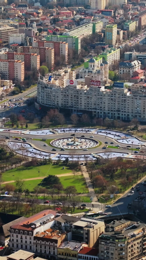 Video - Bucharest, Romania - March 03, 2024: Vertical aerial drone view of the city downtown in sunlight. Roundabout intersection with moving cars, fountains and greenery