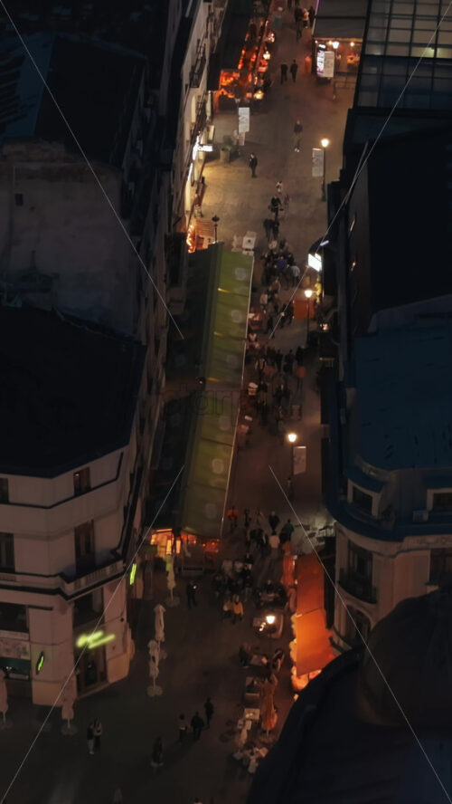 Video - Vertical aerial drone view of walking people among old historical buildings and commercial shops in the old town in the evening. Blue hour in Bucharest, Romania
