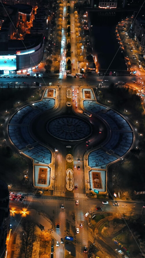 Video - Bucharest, Romania - March 03, 2024: Vertical aerial drone view of a downtown roundabout intersection with moving traffic. Illuminated city in the night
