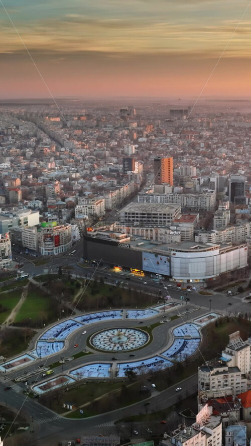 Video - Bucharest, Romania - March 03, 2024: Vertical aerial drone view of the city downtown at sunset. Roundabout intersection with moving cars and fountain. Palace of the Parliament in the distance