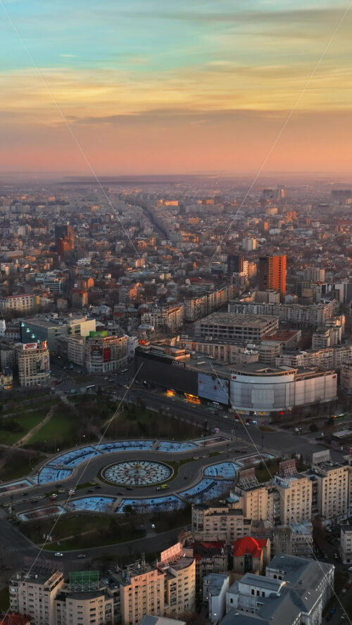 Video - Bucharest, Romania - March 03, 2024: Vertical aerial drone view of the city downtown at sunset. Roundabout intersection with moving cars, fountains and greenery